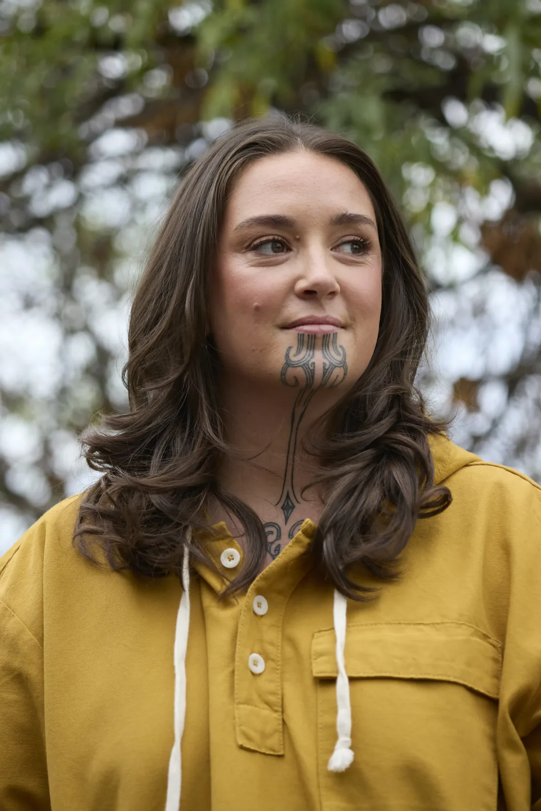 Wahine with a moko kauae in a mustard-yellow hooded shirt, outdoors under a tree, looking off to the side.