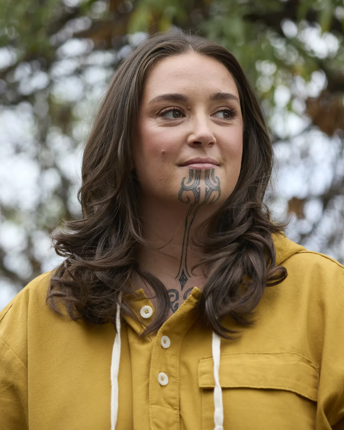 Wahine with a moko kauae in a mustard-yellow hooded shirt, outdoors under a tree, looking off to the side.