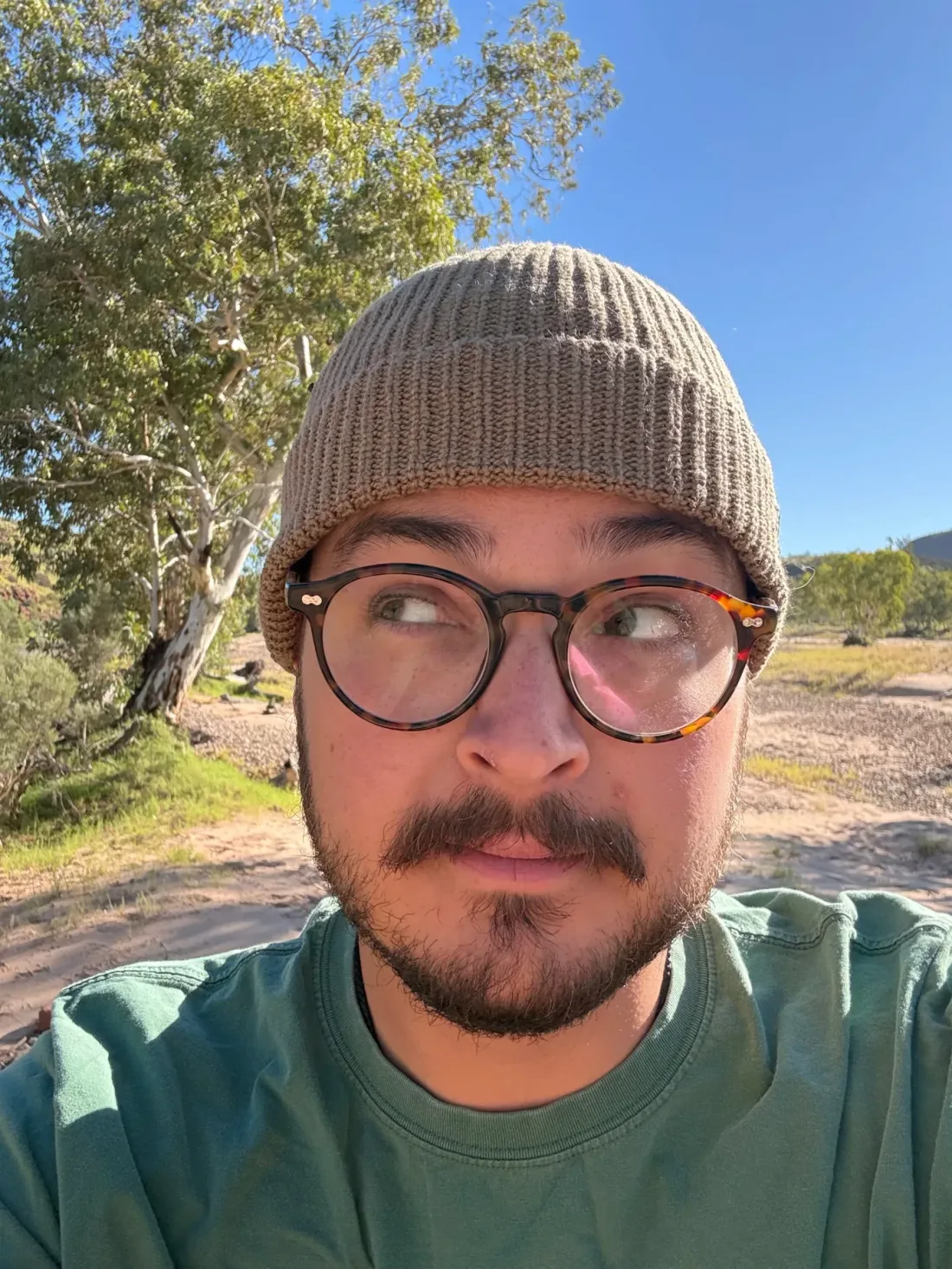 A person in a taupe beanie and round tortoiseshell glasses gives a sceptical side-eye to the camera. Behind them are a white-trunked gum tree and a dry sandy creek bed under a clear blue sky.