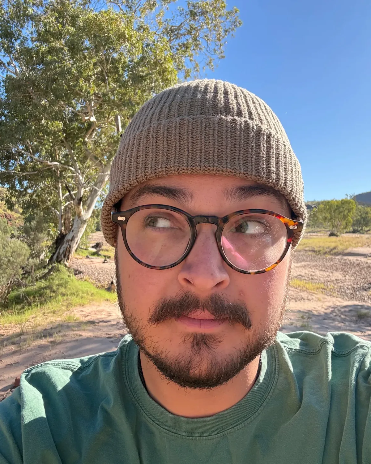 A person in a taupe beanie and round tortoiseshell glasses gives a sceptical side-eye to the camera. Behind them are a white-trunked gum tree and a dry sandy creek bed under a clear blue sky.