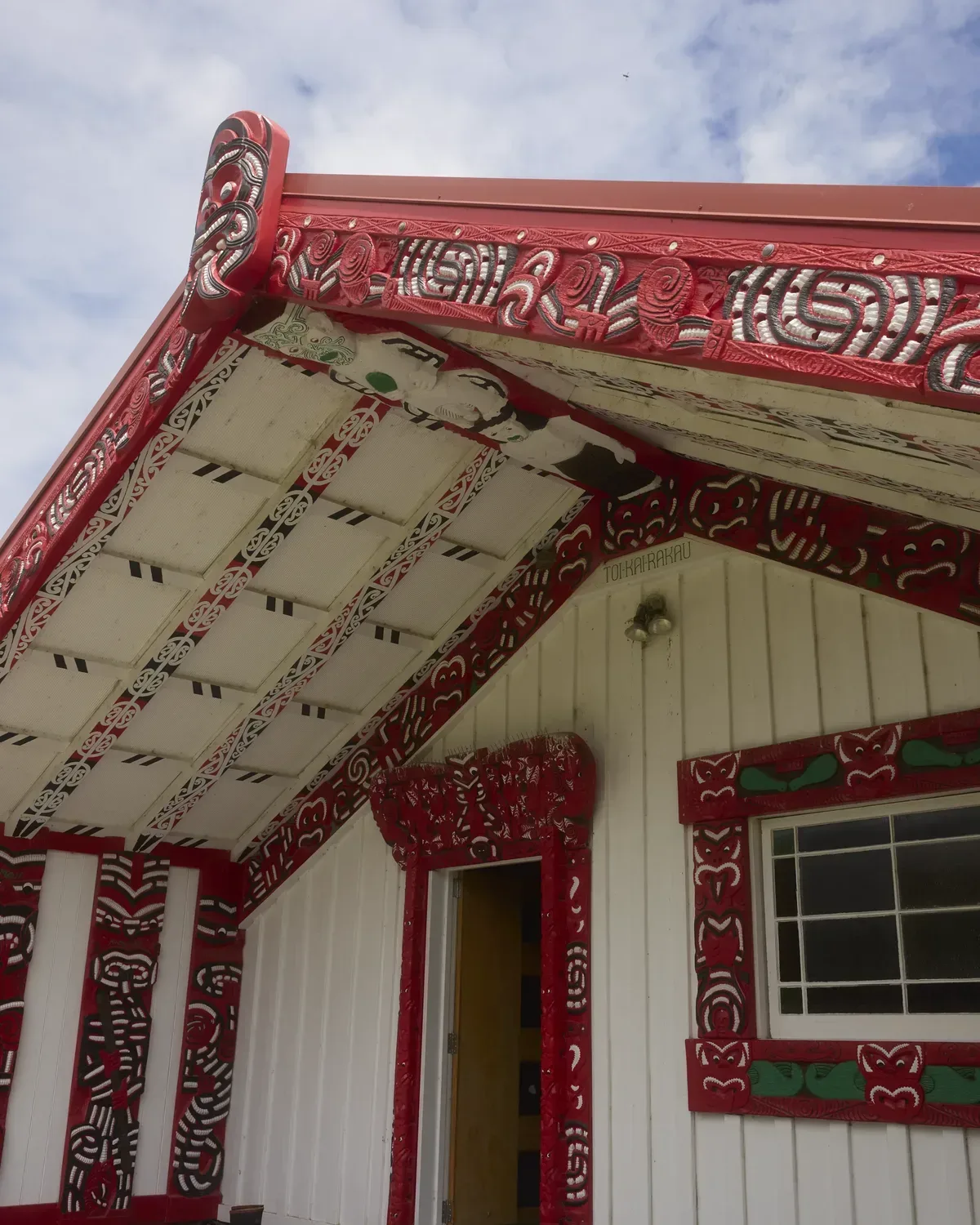 The carved front of a meeting house on a marae, its red carvings and painted rafters under a bright sky.